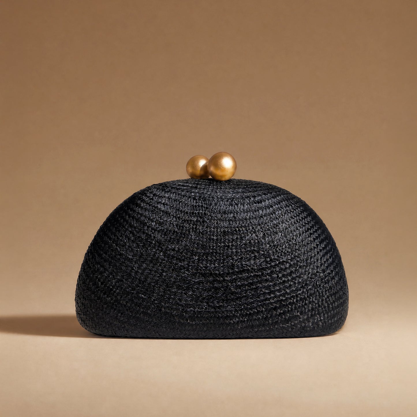 A black woven straw clutch purse with two gold-tone spherical clasp beads on top, set against a solid warm beige background.