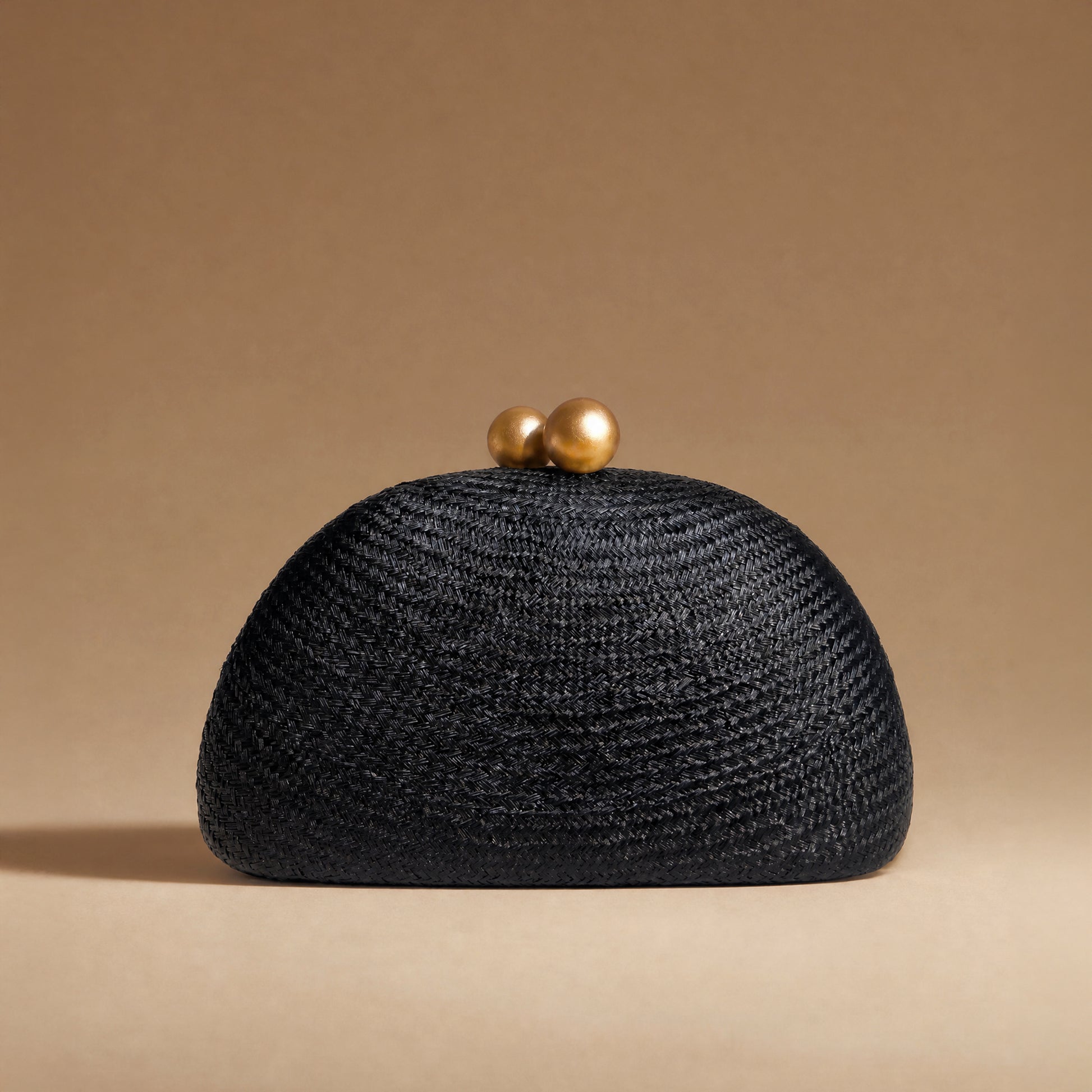 A black woven straw clutch purse with two gold-tone spherical clasp beads on top, set against a solid warm beige background.
