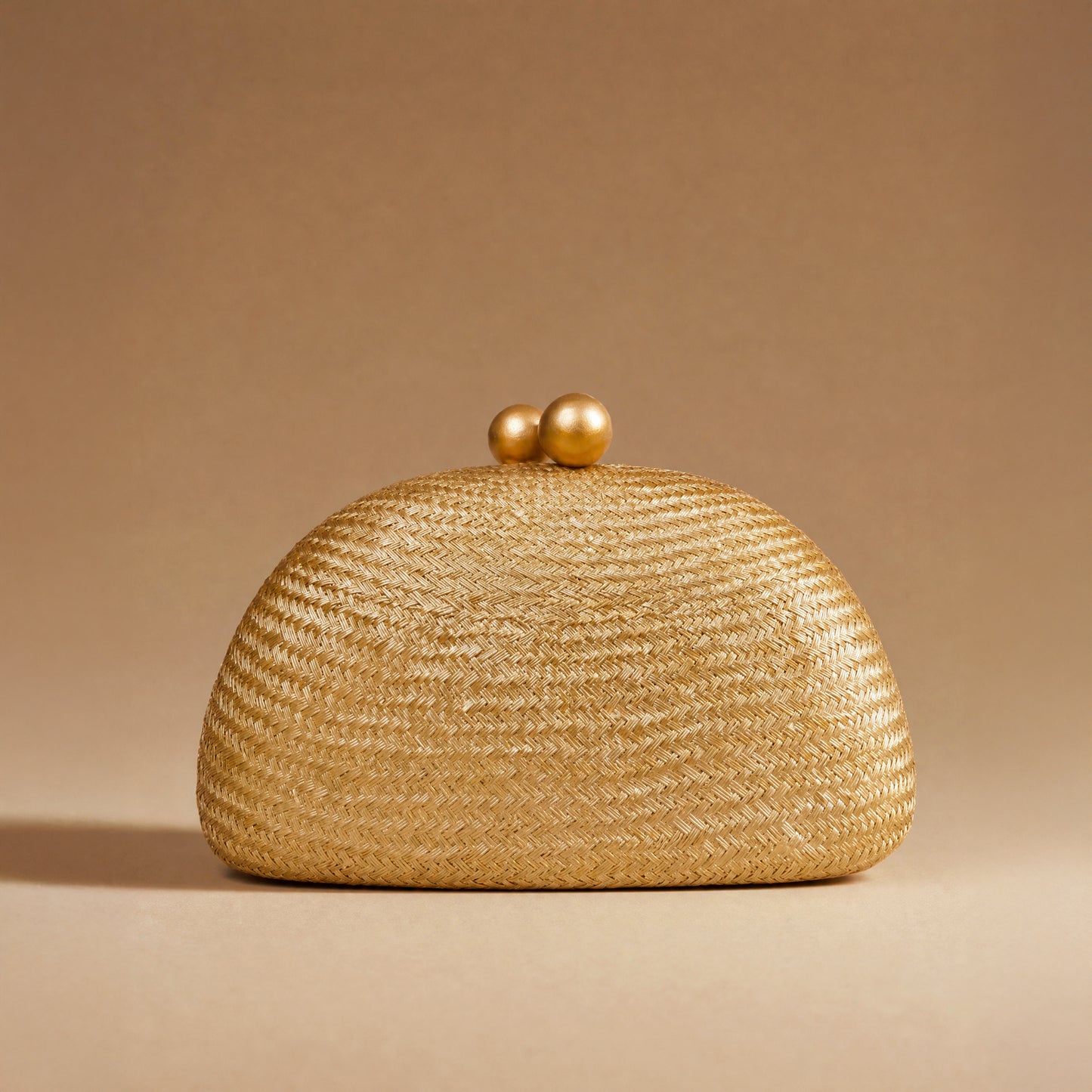 A woven straw clutch purse in a warm golden-beige color, featuring a rounded dome shape and two small wooden ball clasp, set against a solid beige background.
