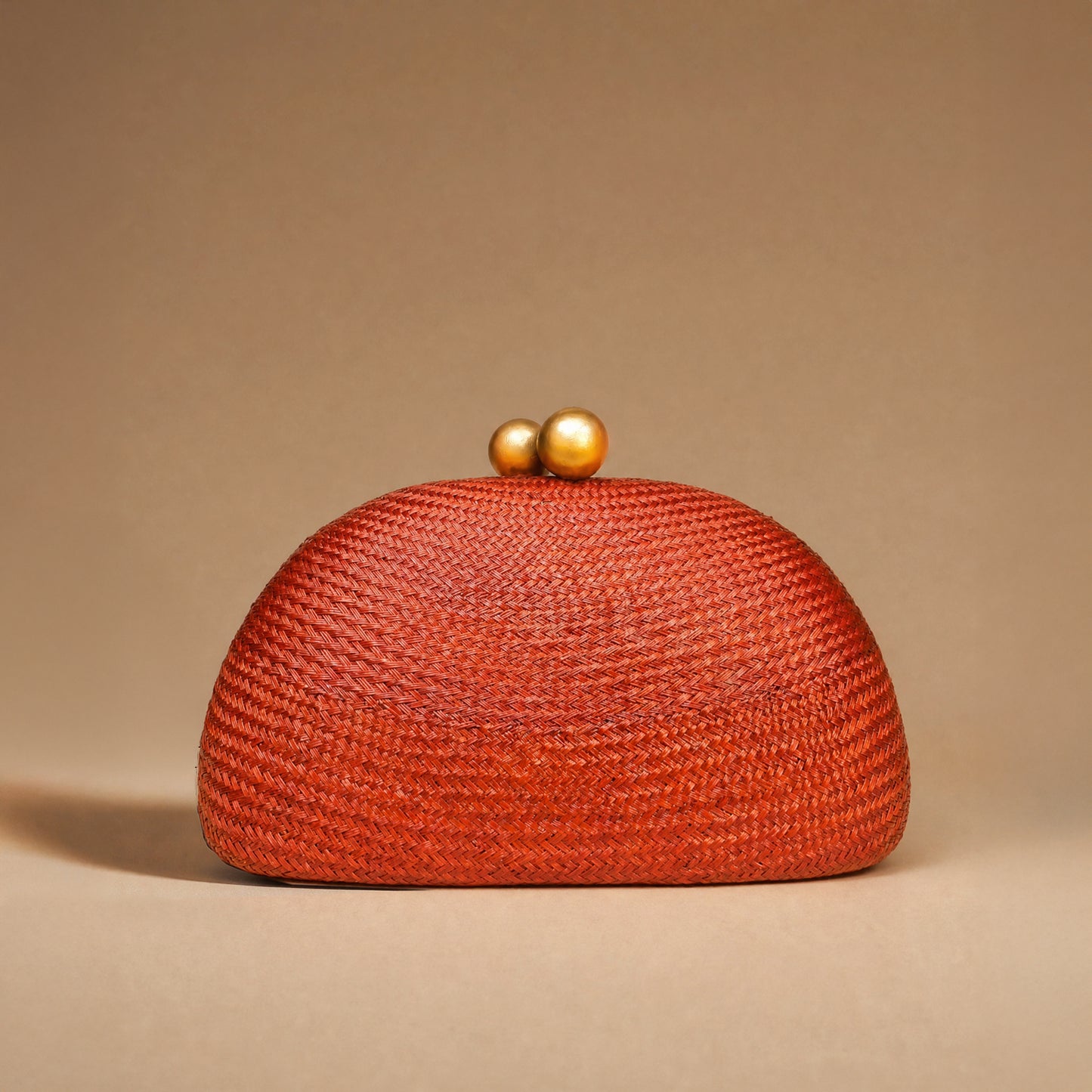 A textured woven clutch bag in a vibrant rust-orange color, featuring a domed shape and two small gold-tone pearl clasp buttons on top, set against a solid warm beige background.