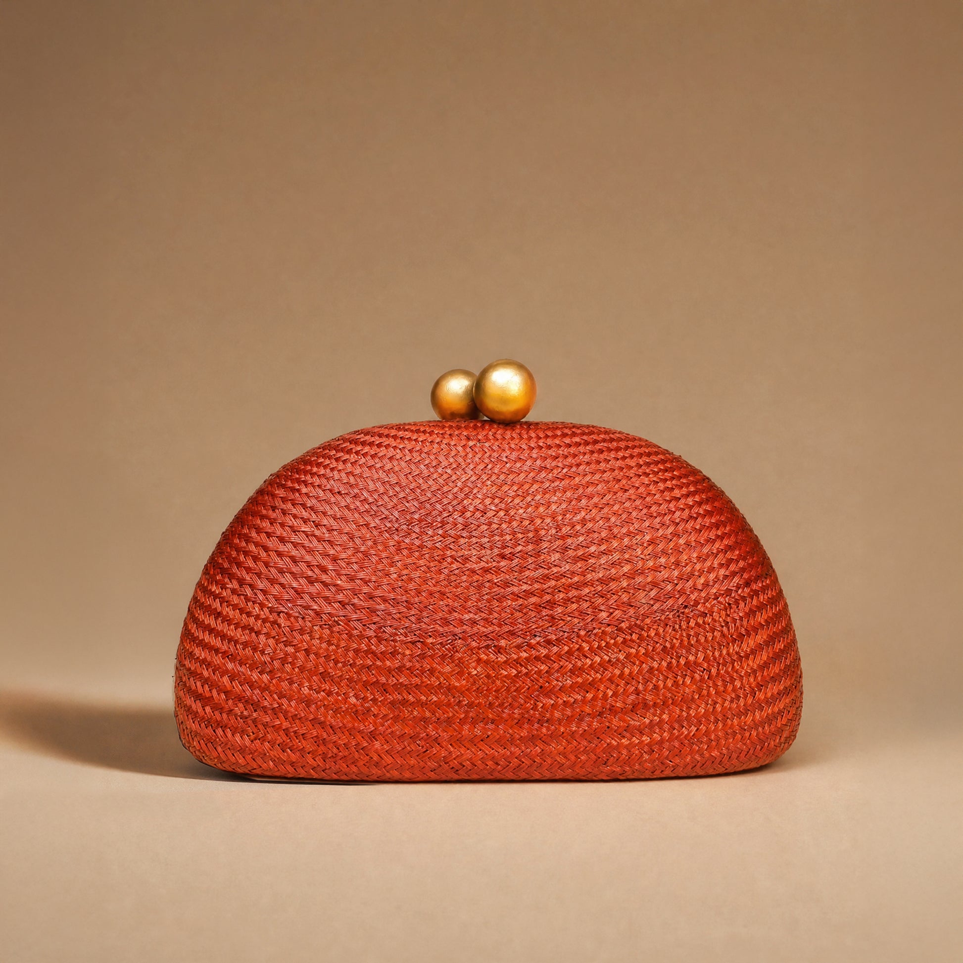 A textured woven clutch bag in a vibrant rust-orange color, featuring a domed shape and two small gold-tone pearl clasp buttons on top, set against a solid warm beige background.