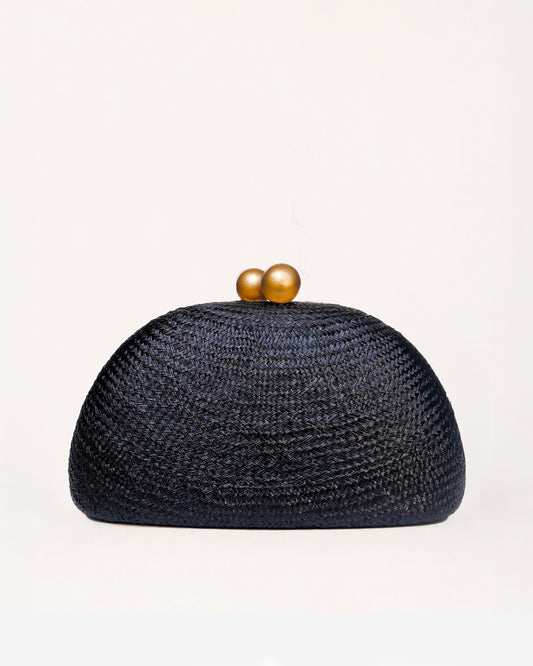 A dark black woven straw clutch bag with a dome shape and two gold wooden balls clasp, set against a plain off-white background.