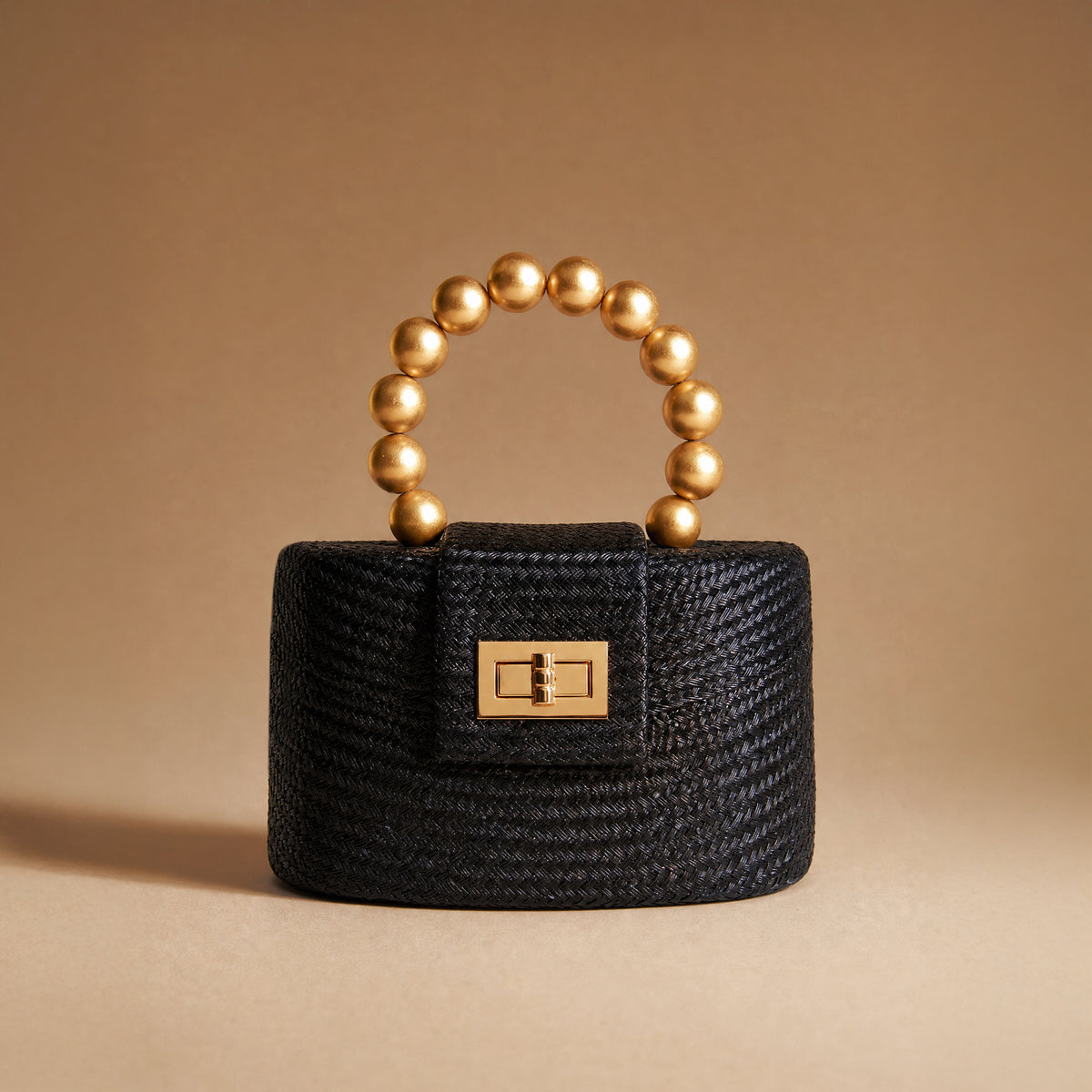 A small structured black woven handbag with a gold-tone clasp and a curved top handle made of metallic gold beads, photographed on a warm beige studio background.