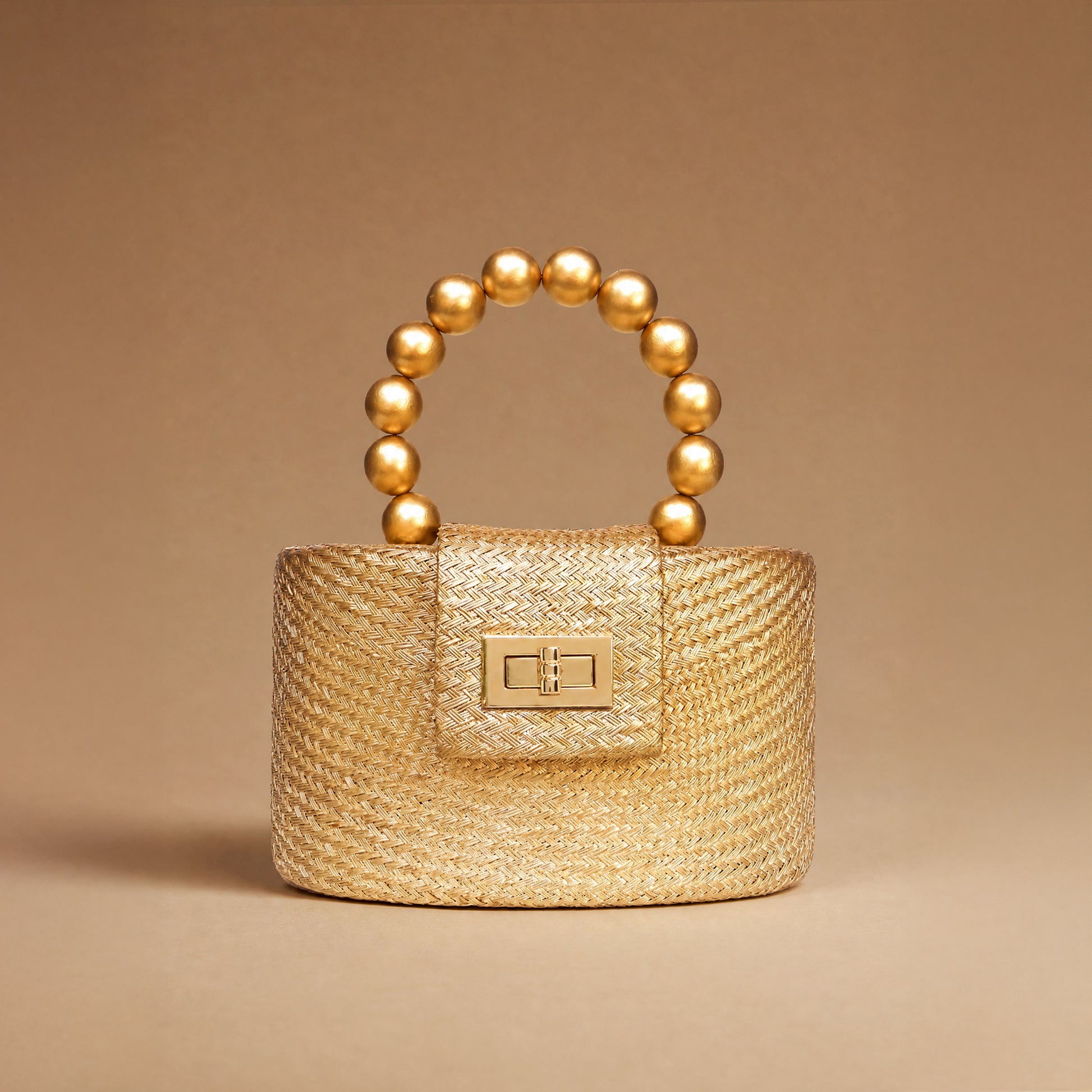 A small structured handbag in woven straw with a gold-tone turn-lock clasp and a curved top handle made of glossy gold beads, photographed on a warm beige studio background.