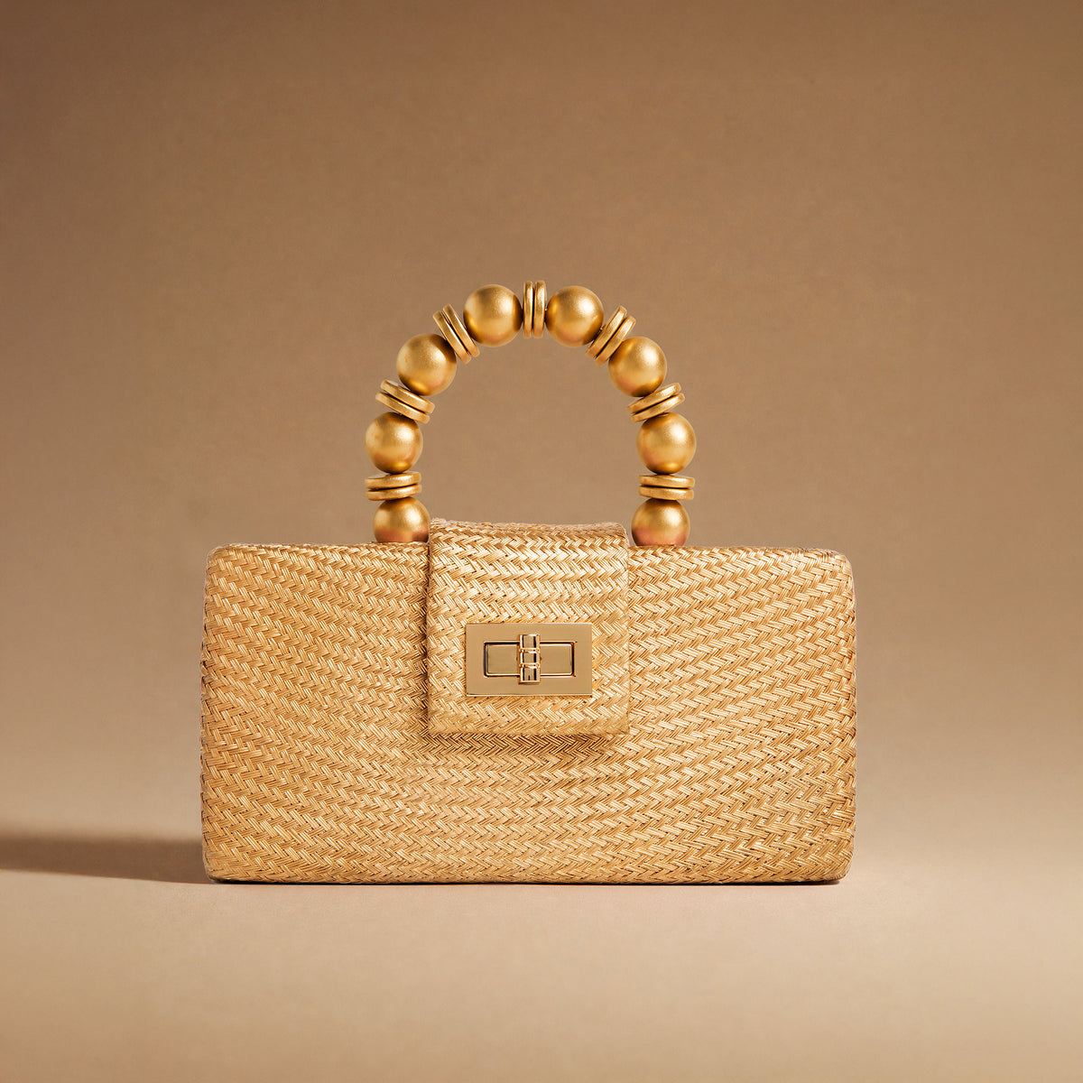 A structured woven straw handbag in a warm gold tone, featuring a curved top handle made of glossy gold-toned beads and a matching gold-toned clasp on the front flap, set against a solid warm brown background.